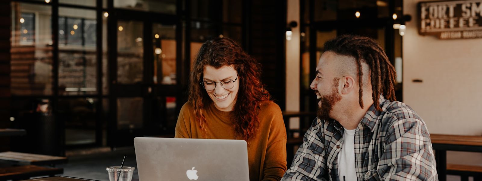 Two professionals at a wooden table with a laptop, engaged in a friendly conversation over coffee, in a modern cafe setting.