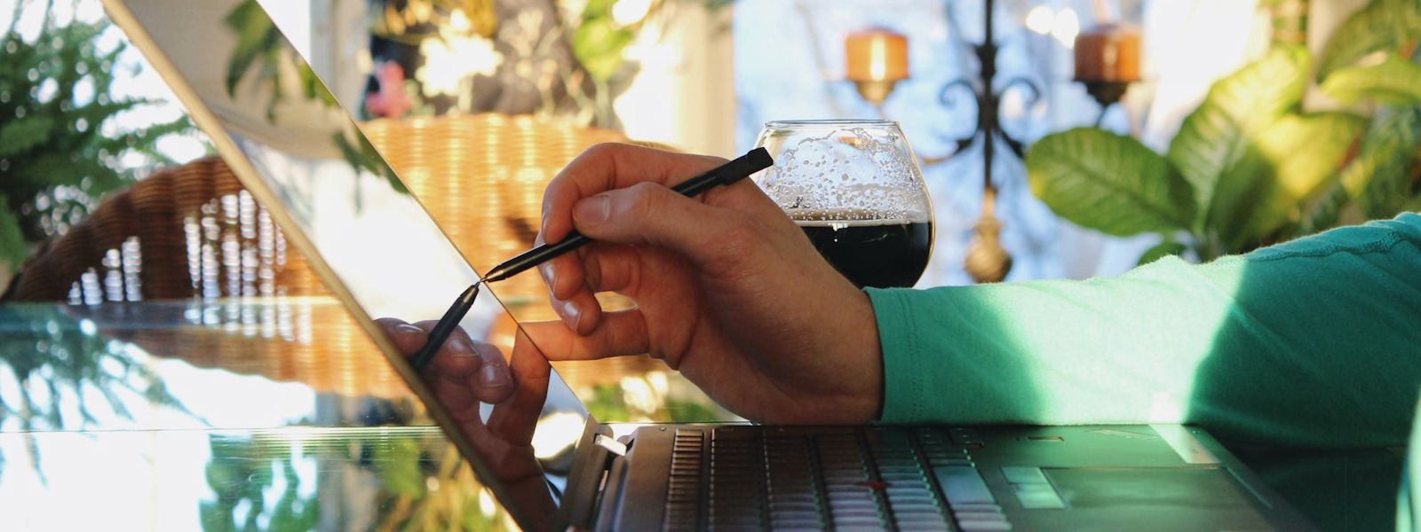 An arm with a green shirt sleeve holding a digital pen to a laptop with a beer next to it in a well-lit room filled with plants.