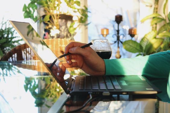 An arm with a green shirt sleeve holding a digital pen to a laptop with a beer next to it in a well-lit room filled with plants.