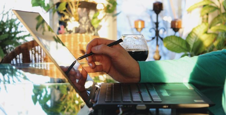 An arm with a green shirt sleeve holding a digital pen to a laptop with a beer next to it in a well-lit room filled with plants.