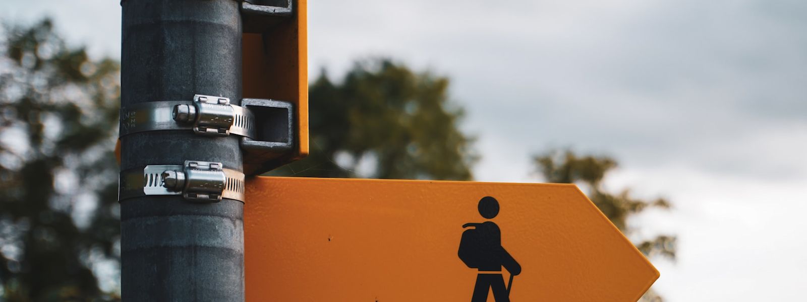 The top of a pole with a yellow sign in the shape of an arrow pointing right, with a picture of a hiking stick figure.