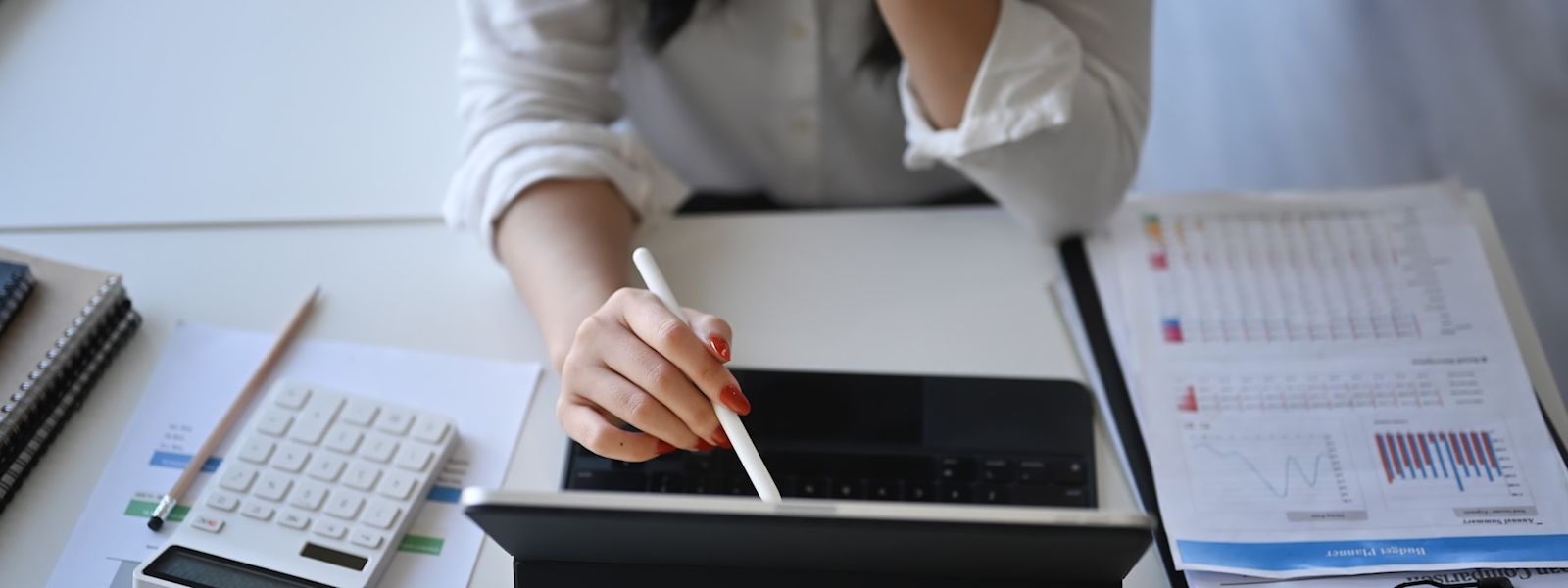 An image of a person using their tablet on a table that has a stack of papers with financial data and graphs.