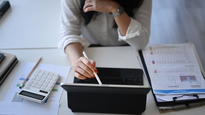 An image of a person using their tablet on a table that has a stack of papers with financial data and graphs.