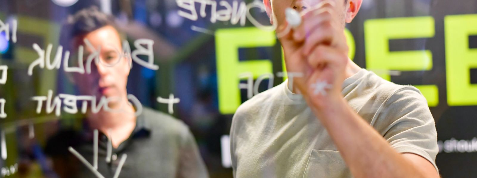 Two people writing on a transparent whiteboard. We're looking at them through the other side of the whiteboard.