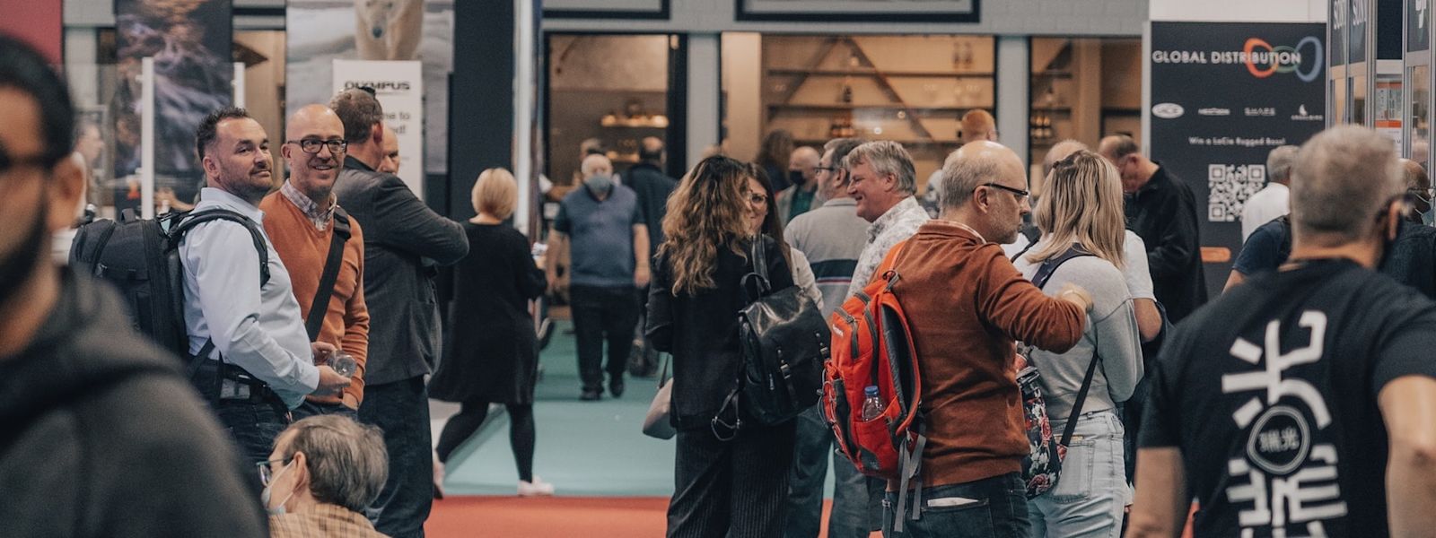 A group of people standing at a convention centre. They're wearing backpacks and are mingling with each other.