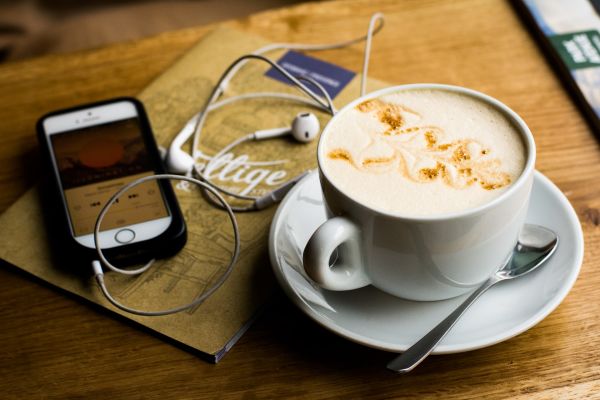On a timber table is a phone playing a podcast with headphones plugged in, next to a coffee.