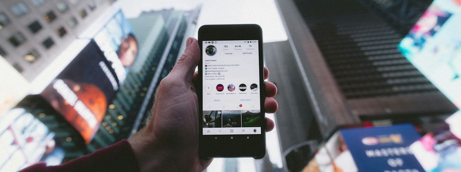 Looking up at a phone screen of an Instagram profile, which draws attention despite crowded Times Square buildings and billboards in the background.