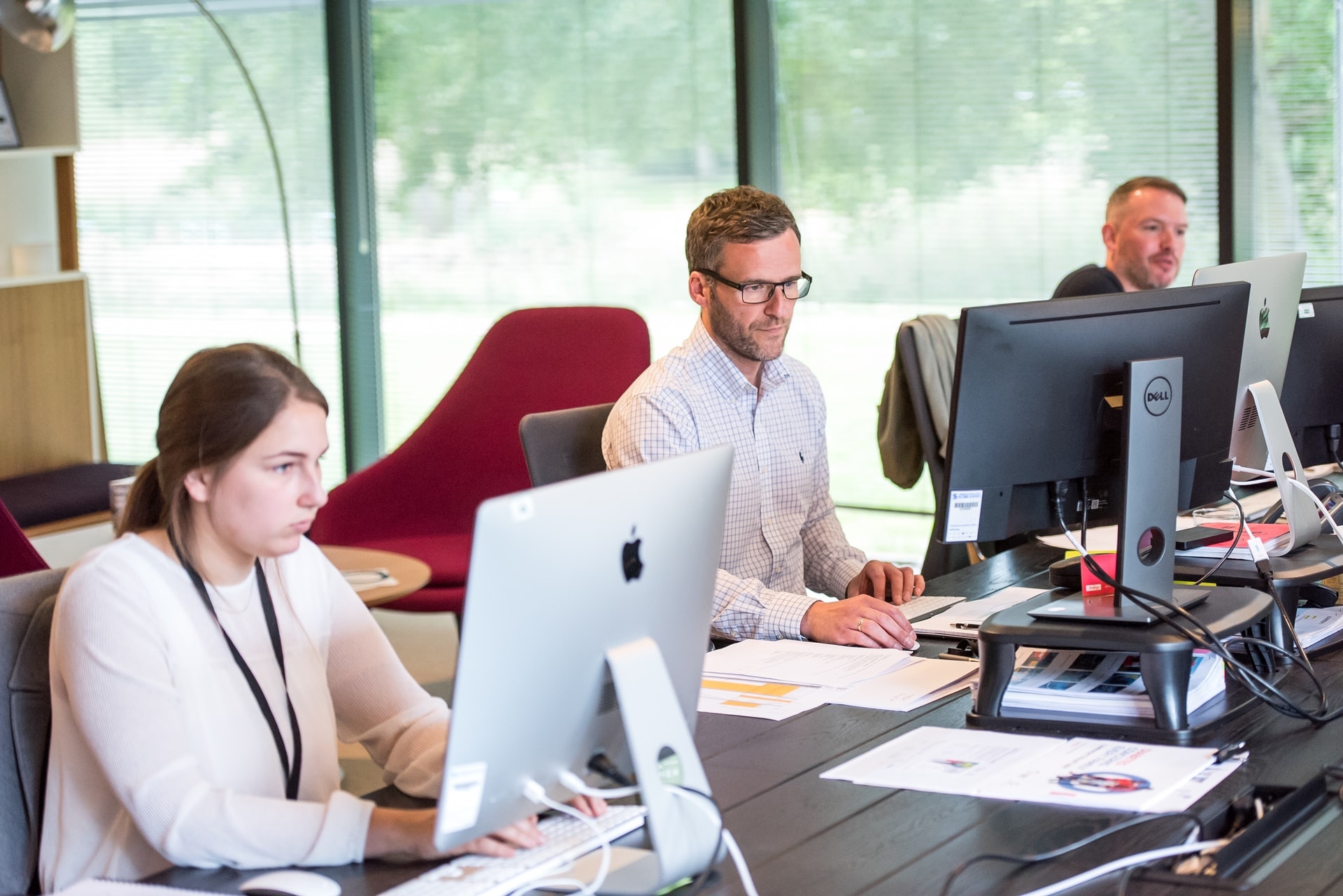 Three people working at a desk, each with their own monitor and papers strewn in front of them.