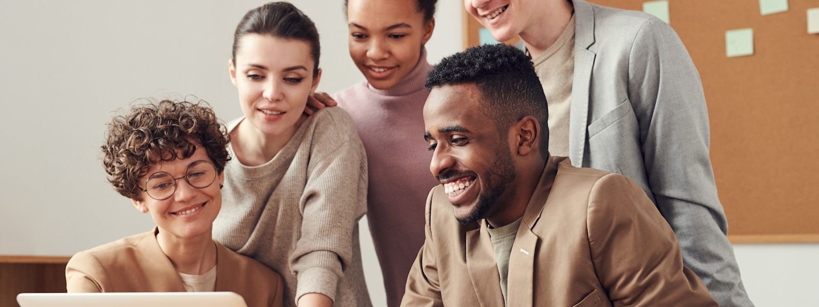 5 young professionals looking at a laptop screen and smiling. There is a pegboard behind them with green stickie notes.