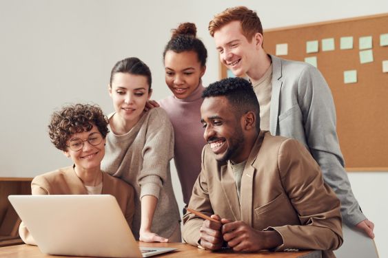 5 young professionals looking at a laptop screen and smiling. There is a pegboard behind them with green stickie notes.