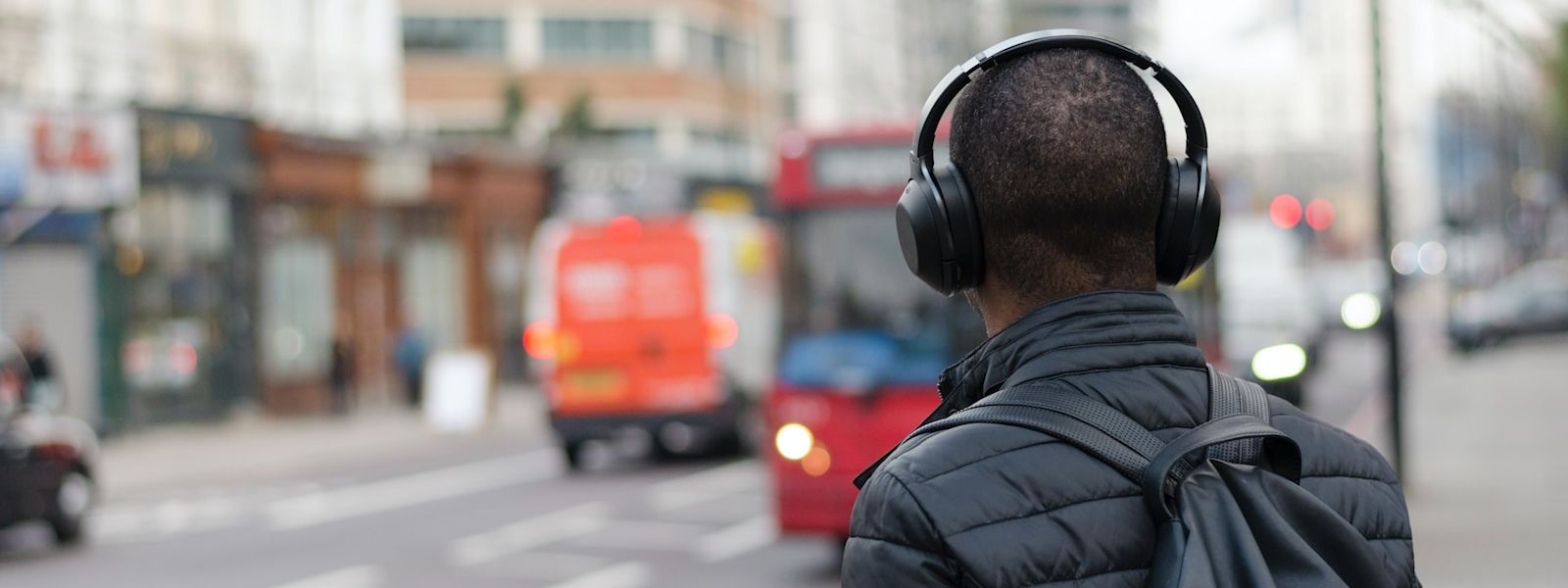 The back of someone standing on the street, looking at a bus while wearing headphones