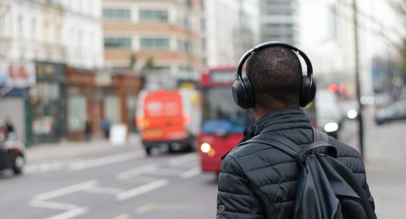 The back of someone standing on the street, looking at a bus while wearing headphones