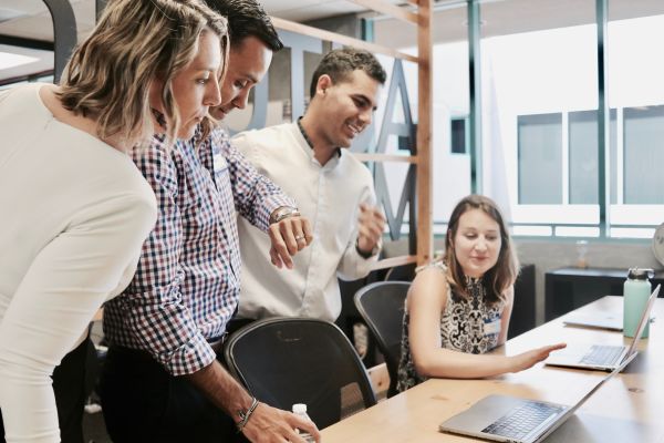 Four people sitting and standing around a desk with laptops on it. They're all looking down at one of the laptop except for one person, whose looking at his watch.