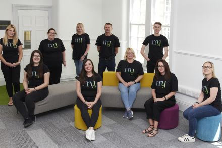 The My Management Accountant team together, sitting and standing around some colorful office furniture. They're all smiling and wearing black tshirts with the My Management Accountant logo.