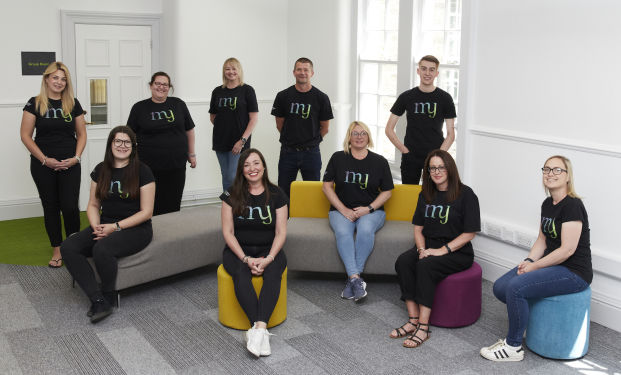 The My Management Accountant team together, sitting and standing around some colorful office furniture. They're all smiling and wearing black tshirts with the My Management Accountant logo.