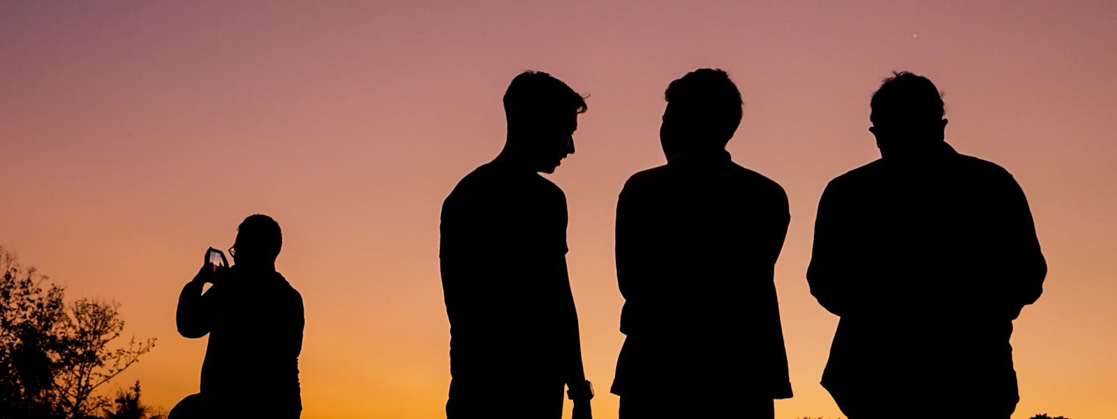4 people looking out over an orangey pink sunset. They are silhouetted. 3 are standing together in the foreground, and the other is in the background on the left, taking a photo on their phone.