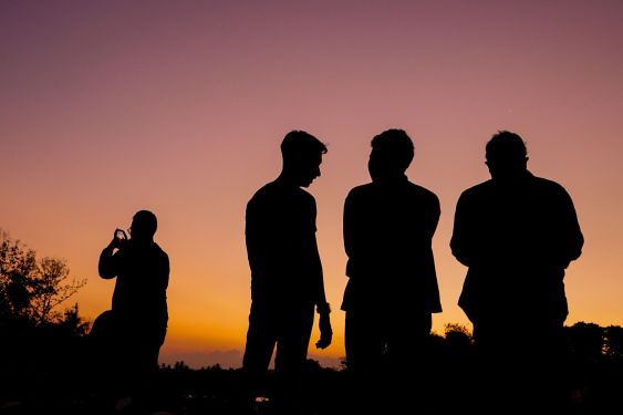 4 people looking out over an orangey pink sunset. They are silhouetted. 3 are standing together in the foreground, and the other is in the background on the left, taking a photo on their phone.