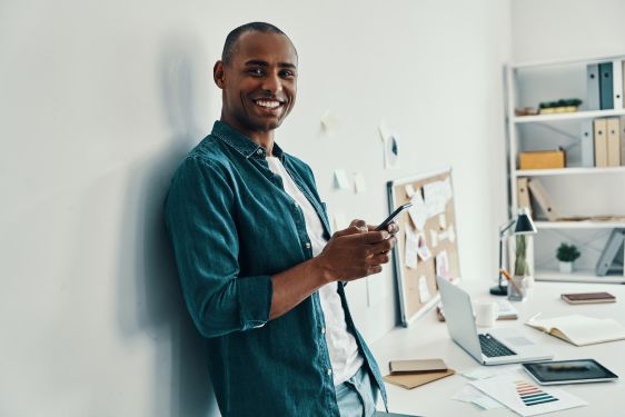 A man is leaning against a wall, smiling at the camera. Behind him is his work desk.