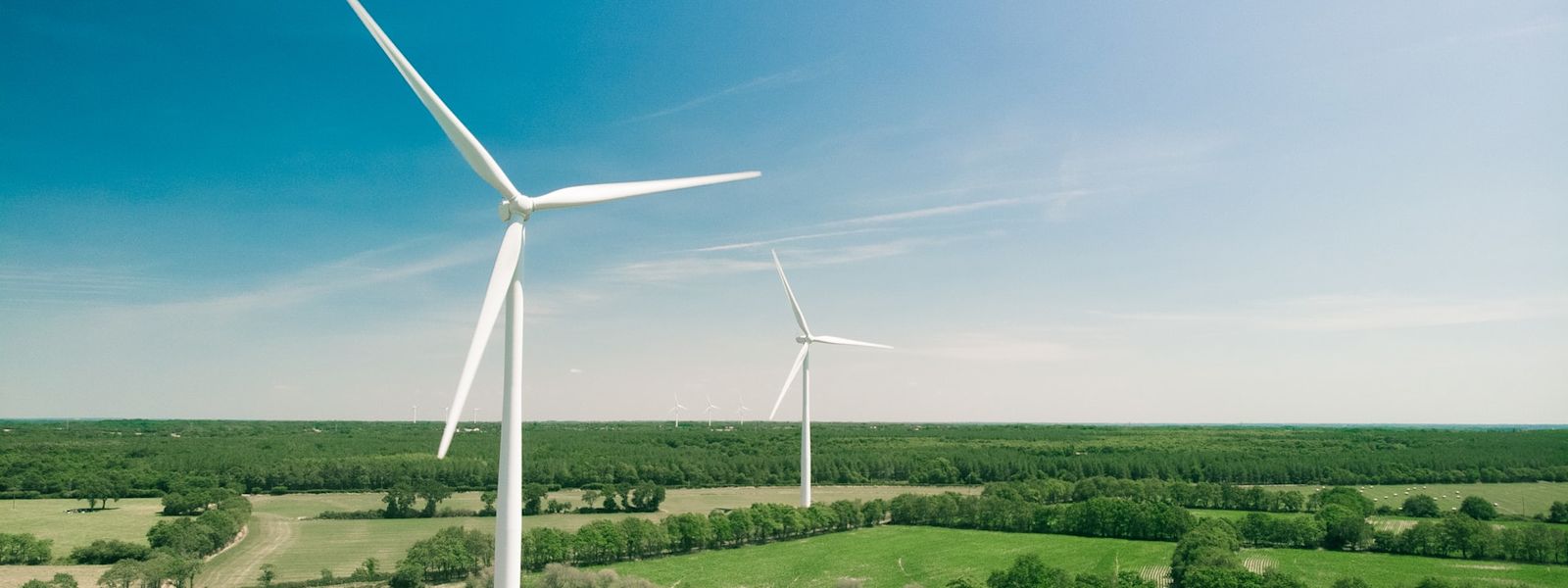 Two wind turbines in a field lined with trees and a blue sky with white, wispy clouds.