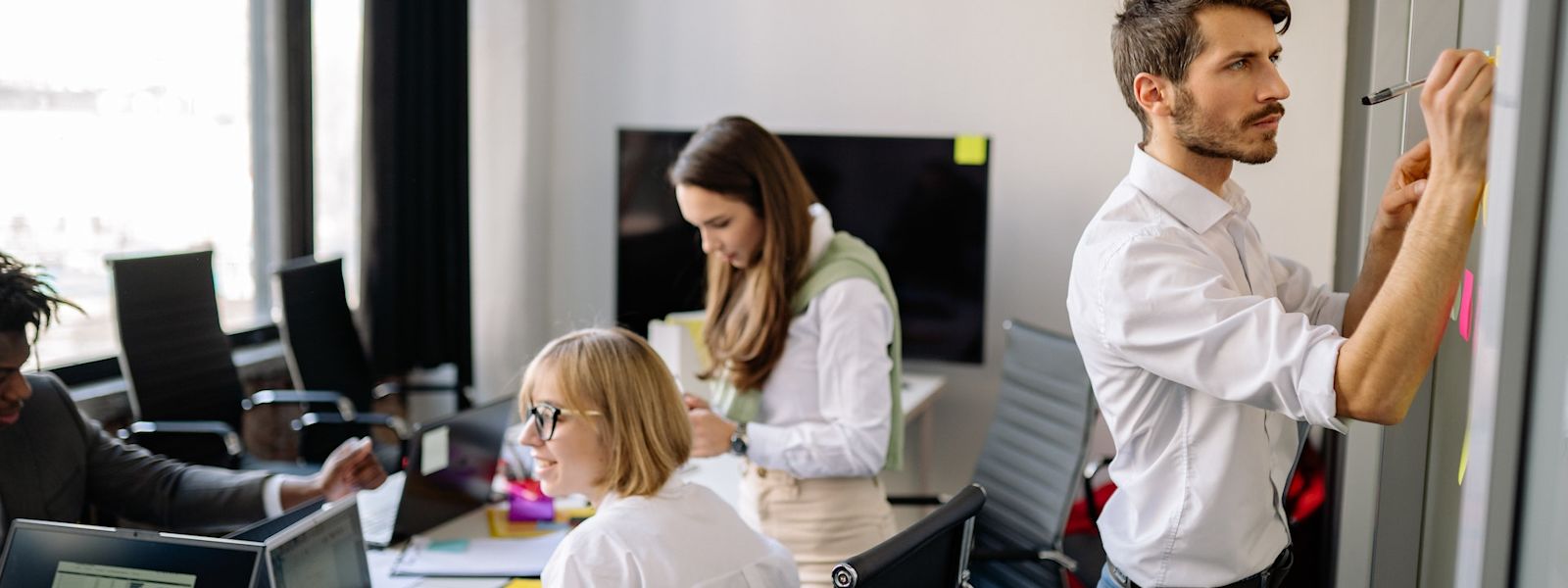 3 people are sitting at a desk covered in paper and laptops, while 1 person is standing up and writing on sticky notes on the wall.