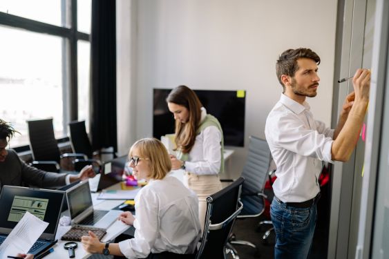 3 people are sitting at a desk covered in paper and laptops, while 1 person is standing up and writing on sticky notes on the wall.