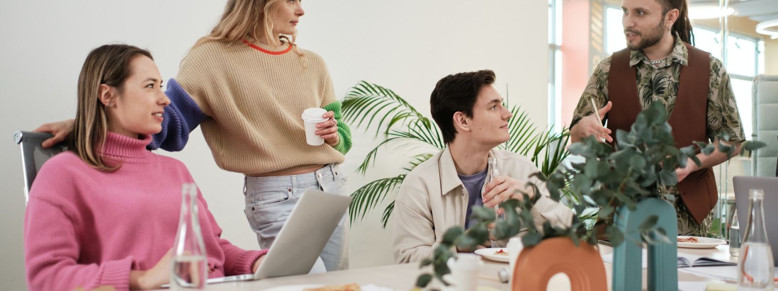 A team of four professionals engaging in a lively discussion at a modern office, with one standing and explaining a point, and the others listening attentively around a table with pizza and water bottles, illustrating collaboration and knowledge sharing.