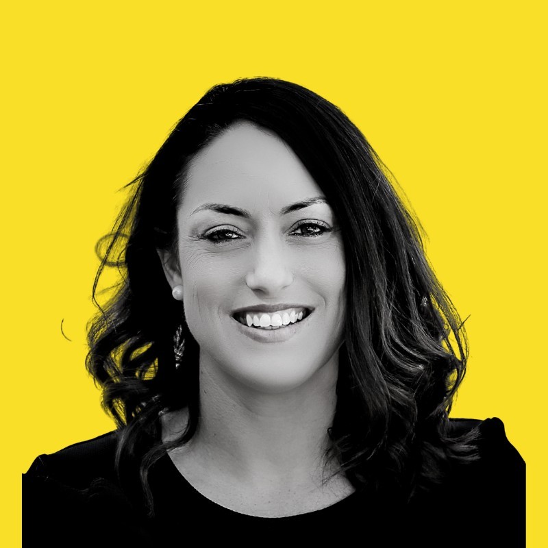Karla Hourigan's headshot. The background is bright yellow, and she has shoulder-length hair and is wearing a black top.