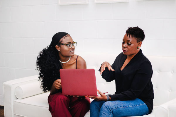 Two women sitting on a couch look at a laptop screen together.