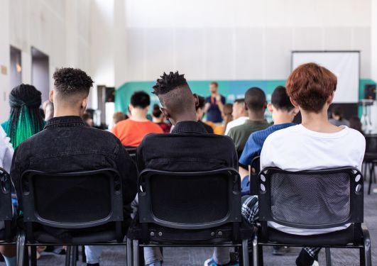 The backs of 3 people sitting and facing a person presenting to a group of people.