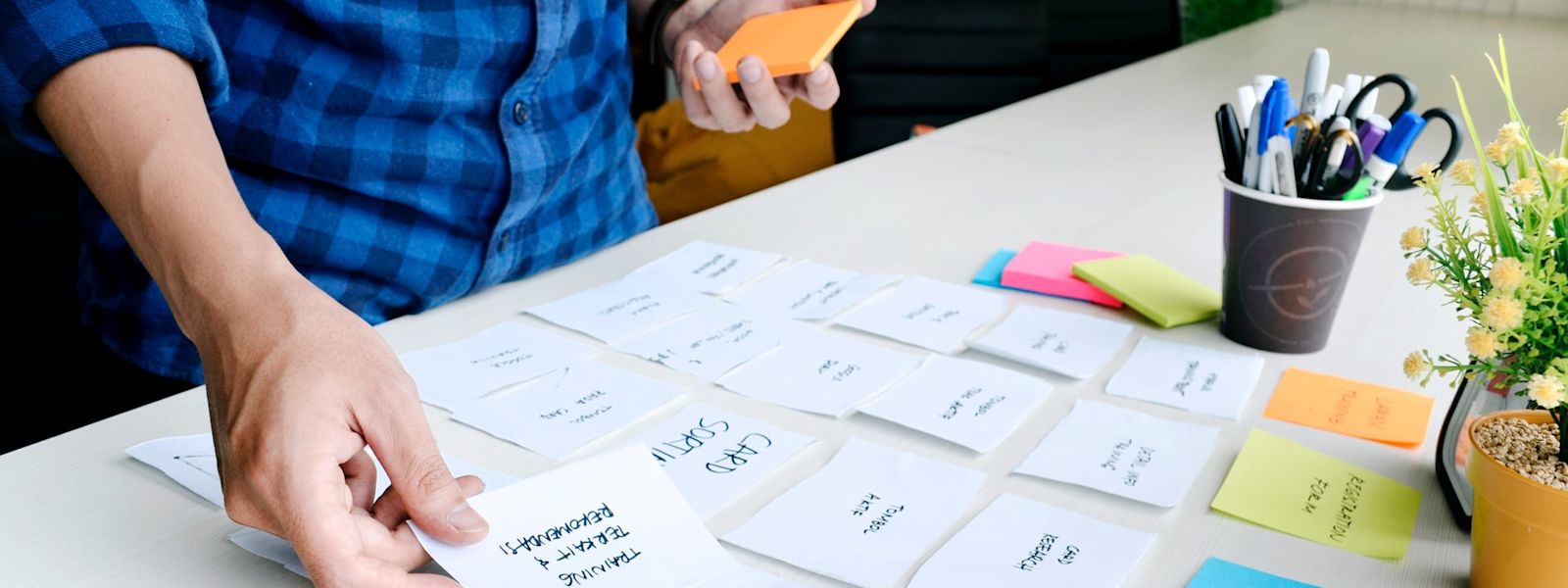 Someone is placing a sticky note on a table top that already has approx. 20 sticky notes on it. The person is wearing a blue checkered shirt and you can only see them from the chest-down.