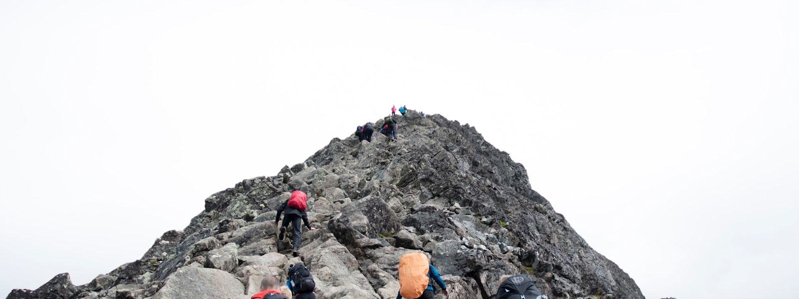 Looking up a rocky mountain from the base. There are 4 people at the base climbing up, each with a colored backpack. At the very peak, there are a handful of other climbers.