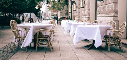 Restaurant with outdoor seating on the street with a brick building and leafy trees in the background