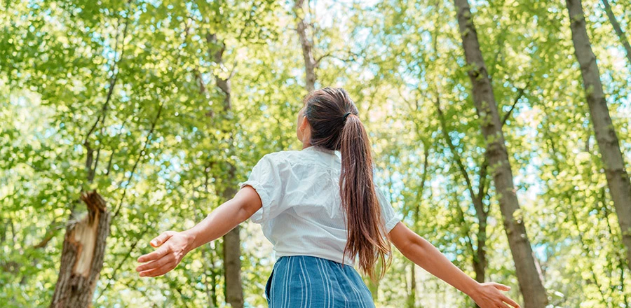 Una chica vista desde atrás con los brazos abiertos en el medio del bosque. Tiene una coleta y lleva una camisa blanca.