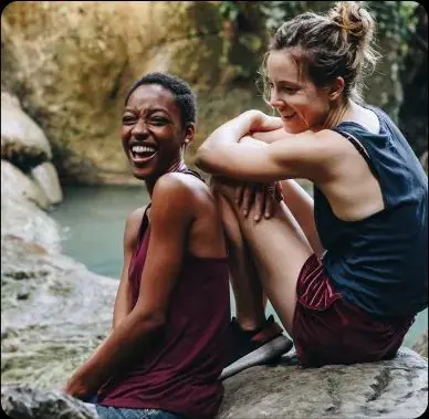 Dos mujeres jóvenes sentadas en una piedra. Una se ríe y la otra la mira y sonríe. Se pueden ver las rocas y el agua al fondo.