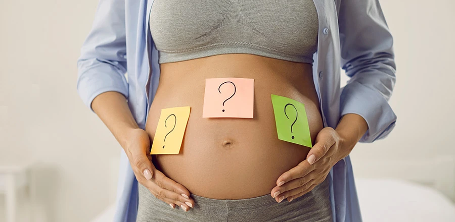 Primer plano del abdomen expuesto de una mujer embarazada. Sobre él hay pegadas tres tarjetas con signos de interrogación.