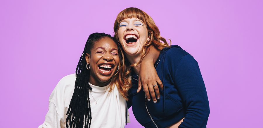 Dos mujeres jóvenes sonriendo están frente a frente hablando y gesticulando. Al fondo se ven árboles y transeúntes.