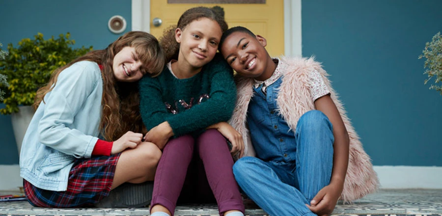 Tres chicas jóvenes sentadas frente a la entrada de una casa con una puerta amarilla. Sonríen y se abrazan.