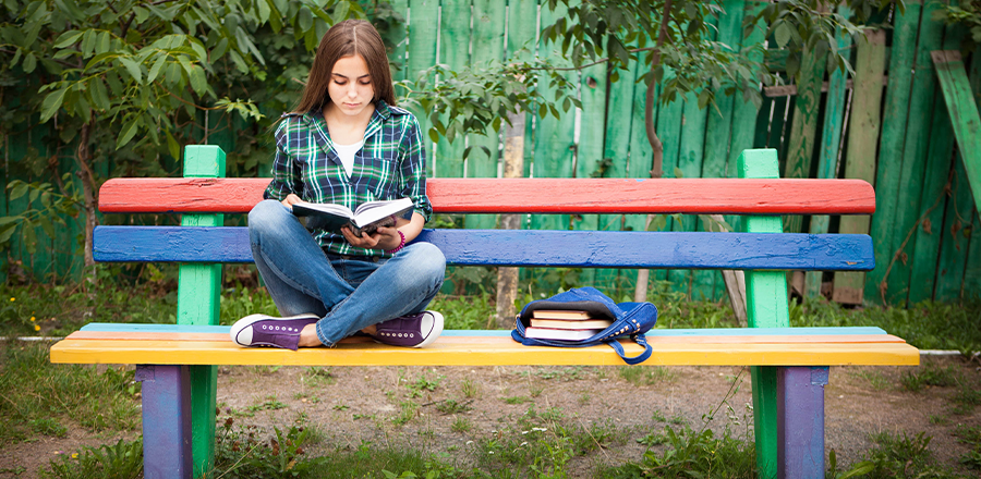Una mujer sentada en un banco leyendo un libro, con una bolsa de libros a su lado