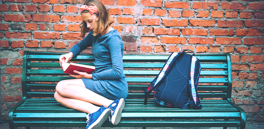 Una mujer leyendo un libro sentada en un banco, con su mochila a su lado