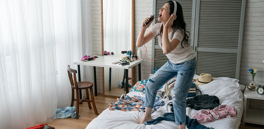 Una mujer de pie sobre la cama con los auriculares puestos y cantando con el mando a distancia en la mano