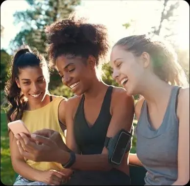 Tres chicas sentadas al aire libre. Una de ellas sostiene un teléfono y muestra algo. Todas miran y sonríen.