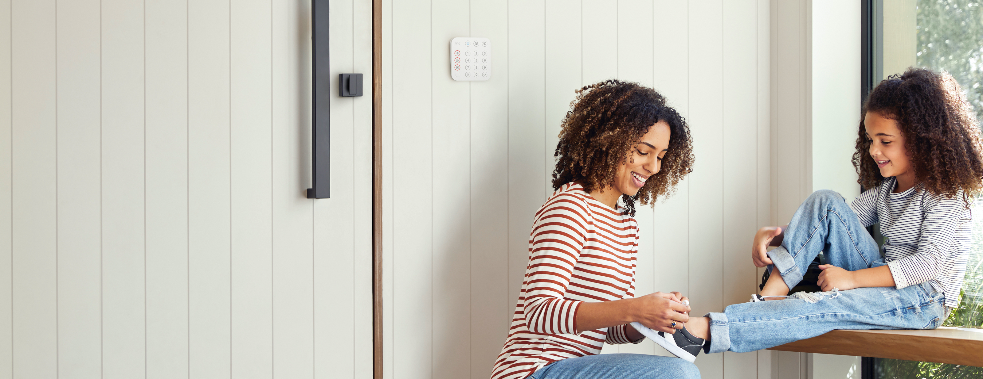 Lächelnde Mutter bindet Tochter die Schuhe nahe der Tür, Ring Alarm Keypad an der Wand dahinter.