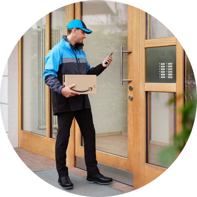 Corriere Amazon con pacco e smartphone davanti all'ingresso di un condominio con citofono.
