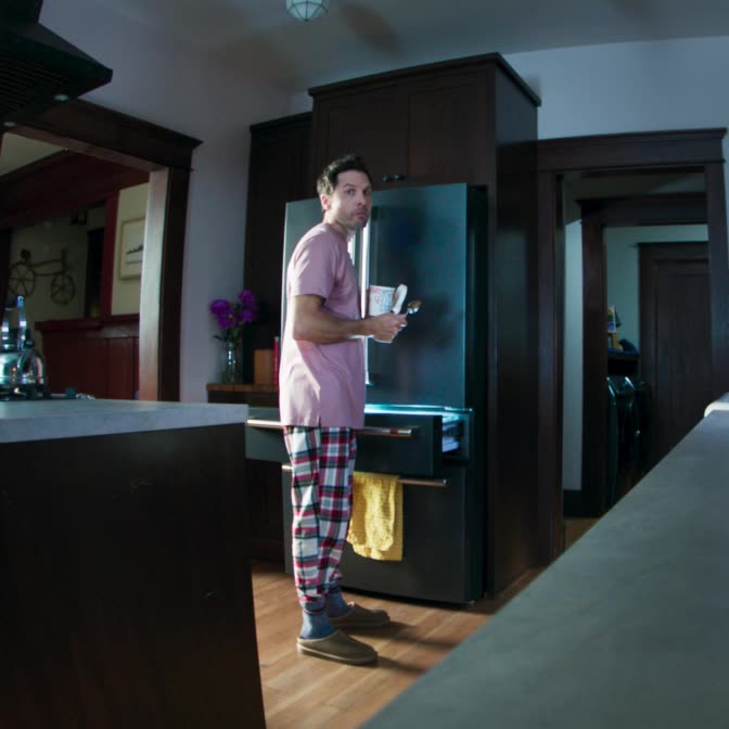 Image shows the view of a home kitchen through an indoor security camera, showing a man in pajamas standing by a refrigerator eating ice cream at night. 