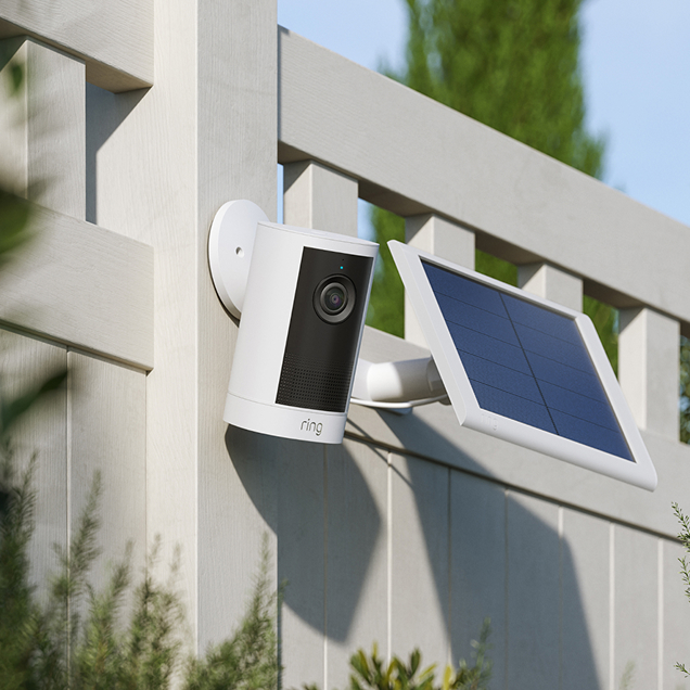Ring Outdoor Camera 4K Plus with Solar Panel mounted on a white fence, surrounded by green foliage.