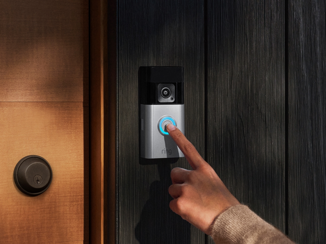 Person pressing illuminated button on Ring Video Doorbell mounted on dark wooden doorframe near a modern door handle.