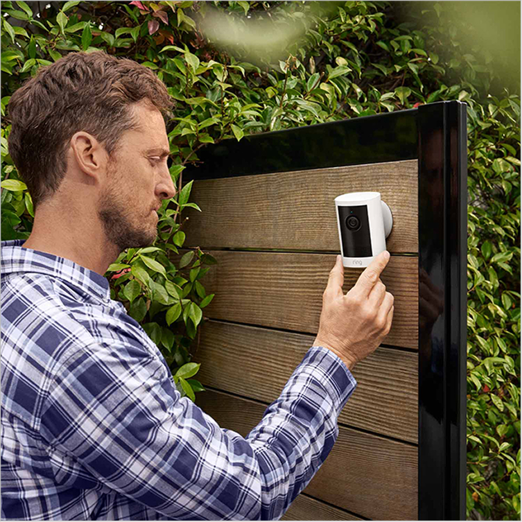 Person in garden installing an Outdoor Camera (2nd Gen) on a fence with foliage in the background, demonstrating easy install mount