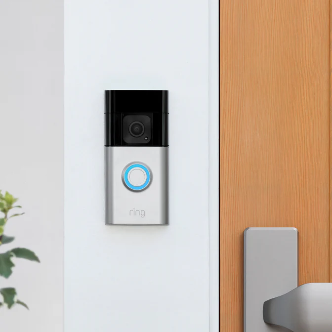 Ring Battery Doorbell Plus mit Kamera und beleuchtetem Knopf neben einer Holz-Haustür montiert.