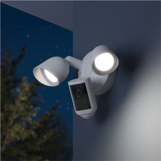 Floodlight Cam (2e génération) blanche, la nuit, installée sur le côté d'une maison, les deux projecteurs allumés.