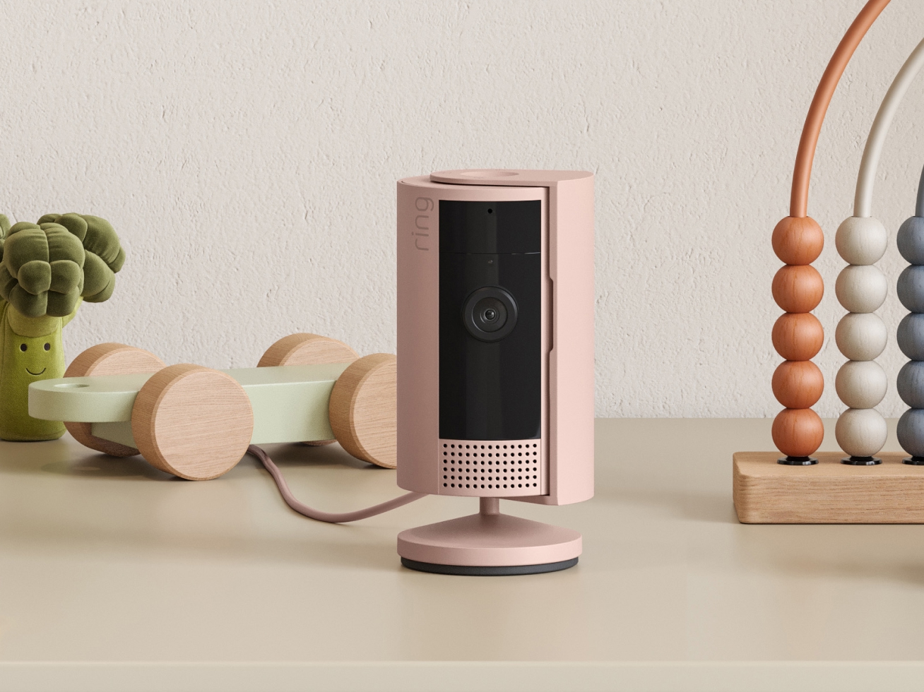 Blush pink Ring indoor security camera on tabletop beside wooden toys, including an abacus and toy broccoli.